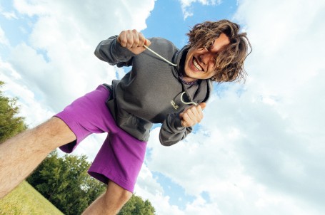 Frau mit lockigem Haar und einem orangen T-Shirt hockt auf einem Felsen vor einer bergigen Landschaft.