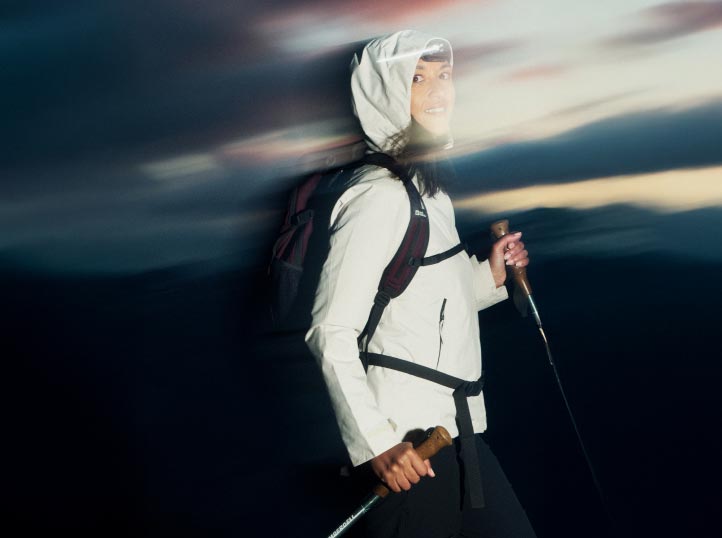 Eine Person in Wanderkleidung, einschließlich weißer Jacke und Kapuze, lächelt, während sie Trekkingstöcke hält. Der verschwommene Dämmerungshimmel suggeriert Bewegung und Abenteuer.
