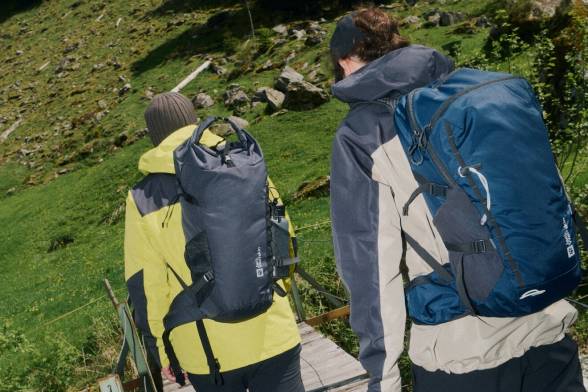 Zwei Wanderer von hinten mit Rucksäcken auf dem Rücken laufen über eine Holzbrücke