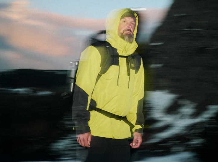 Mann in leuchtend gelber Jacke und Rucksack beim Wandern in der Abenddämmerung, Bewegungsunschärfe verleiht dem Bild Dynamik.