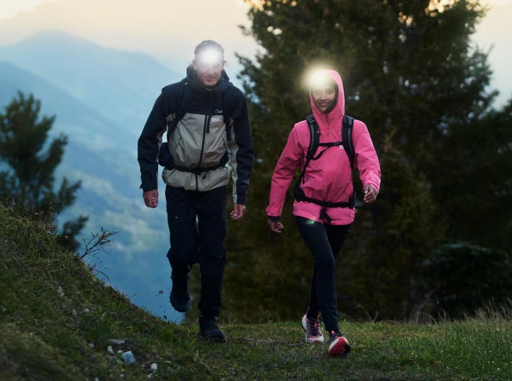Zwei Wanderer mit Stirnlampen gehen in der Dämmerung auf einem Waldweg. Sie tragen Jacken, eine schwarz und grau, die andere rosa.